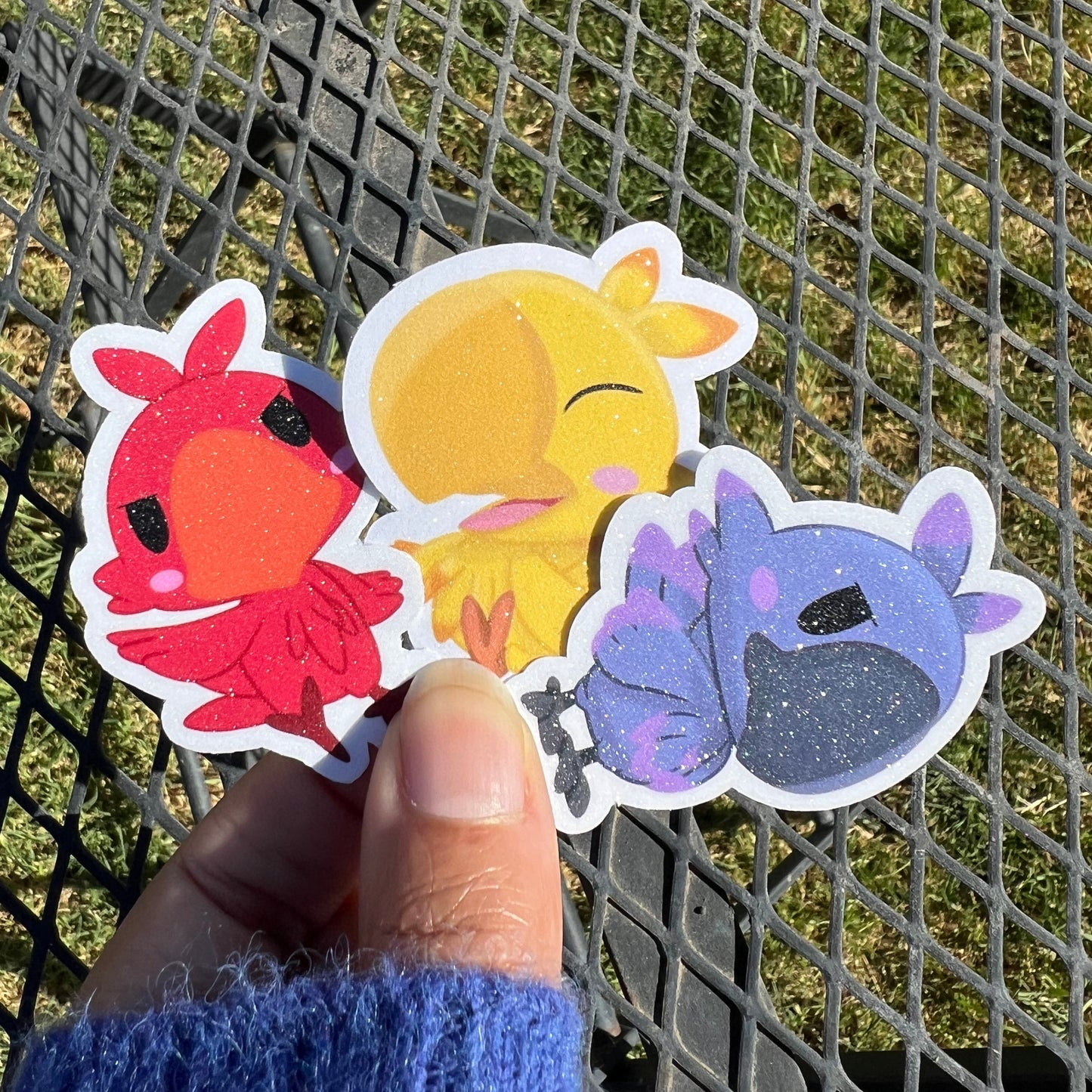 Three colorful stickers of a butterfly, sun, and flower held by a hand against a metal fence background.