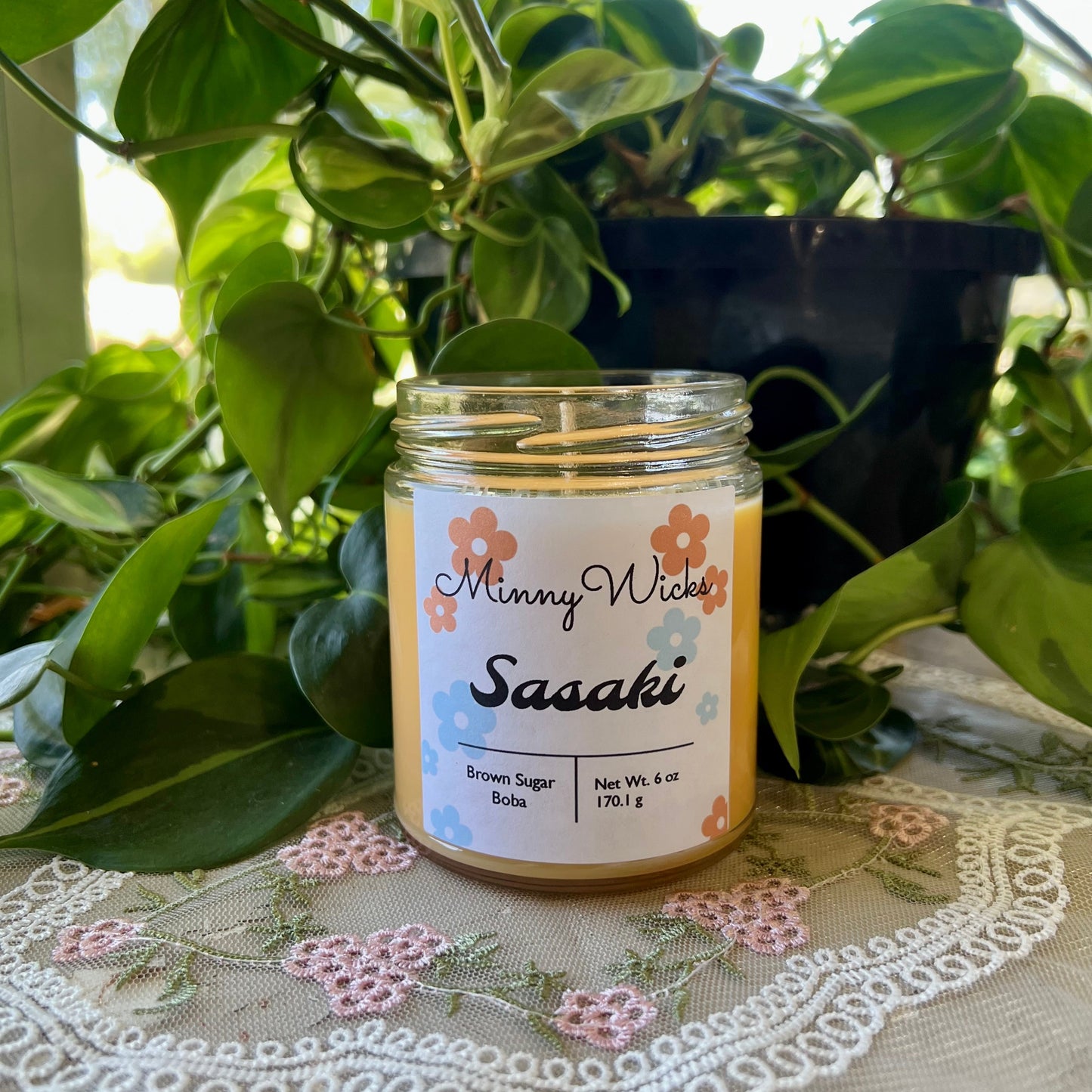 Candle jar labeled 'Sasaki' by 'Minny Wicks' on a table with plants in the background