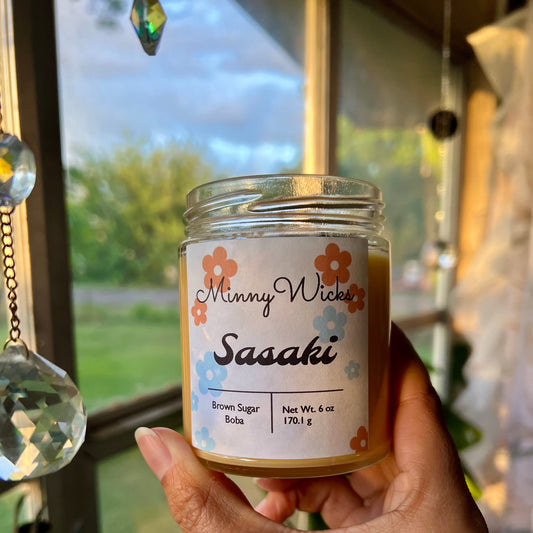 Hand holding a jar of 'MinnyWicks Sasaki' brown sugar boba candle in front of a window with a view of greenery.
