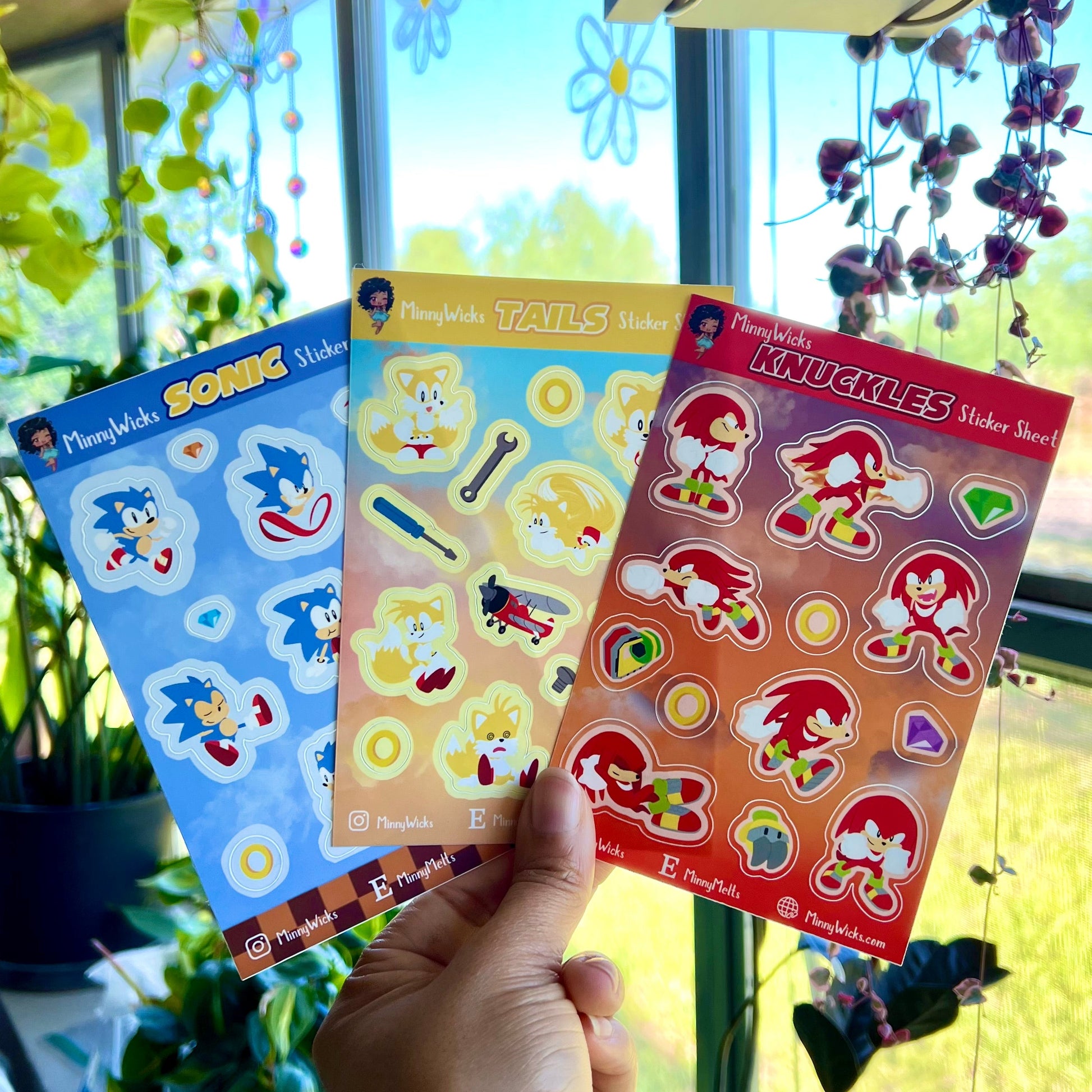 Three sonic sticker sheets held by a hand in front of a window with plants