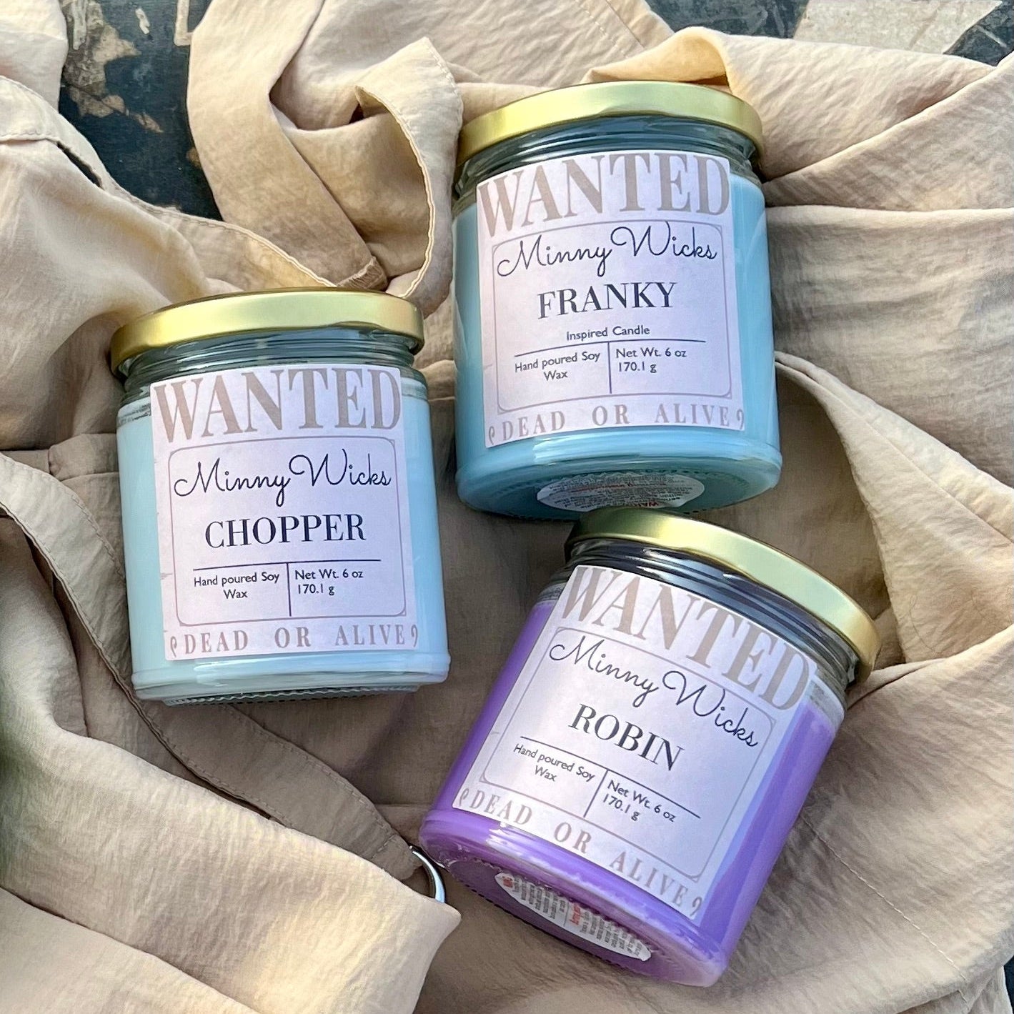Three 'Wanted' Minny Wicks candles on a textured fabric background
